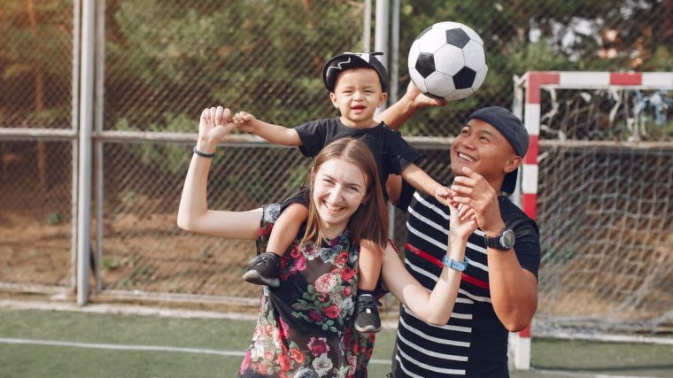 Positive smiling diverse parents giving piggyback ride to adorable toddler son while spending weekend together with football ball on sports ground on sunny day