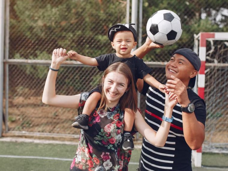Positive smiling diverse parents giving piggyback ride to adorable toddler son while spending weekend together with football ball on sports ground on sunny day